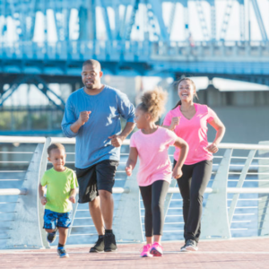 A young black family with two small children staying fit together. They are power walking along the waterfront of a city on a sunny day, having fun with the 7 year old little girl and her 3 year old brother leading the way.